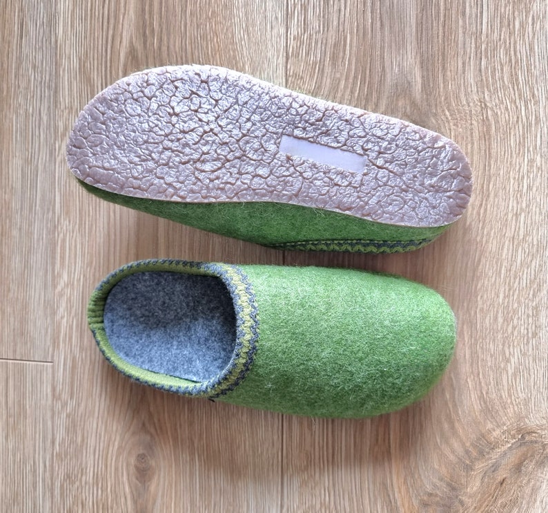 Puede incluir: Un par de zapatillas de fieltro verde con interior gris y suela marr&oacute;n texturizada. Las zapatillas tienen un ribete verde oscuro y gris alrededor de la abertura. Las zapatillas se muestran sobre una superficie de madera marr&oacute;n claro.