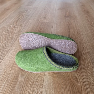 Puede incluir: Un par de zapatillas de fieltro verde con interior gris y suela marr&oacute;n. Las zapatillas tienen un ribete cosido en verde oscuro y gris. Las zapatillas se muestran sobre un suelo de madera.