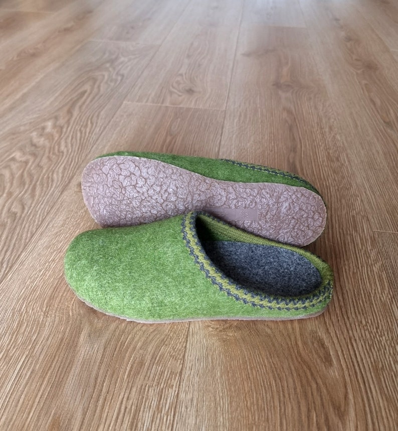 Puede incluir: Un par de zapatillas de fieltro verde con interior gris y suela marr&oacute;n. Las zapatillas tienen un ribete cosido en verde oscuro y gris. Las zapatillas se muestran sobre un suelo de madera.