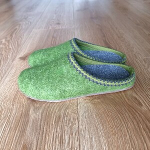Puede incluir: Un par de zapatillas de fieltro verde con ribete verde claro y gris. El interior es gris. Las zapatillas est&aacute;n sobre un suelo de madera.