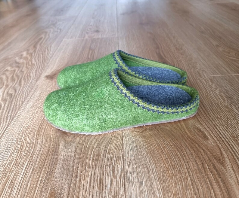 Puede incluir: Un par de zapatillas de fieltro verde con ribete verde claro y gris. El interior es gris. Las zapatillas est&aacute;n sobre un suelo de madera.