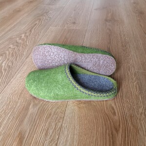 Puede incluir: Un par de zapatillas de fieltro verde con interiores y suelas grises. Las zapatillas tienen un ribete cosido en un color contrastante. Una zapatilla est&aacute; en posici&oacute;n vertical y la otra al rev&eacute;s, mostrando la suela.