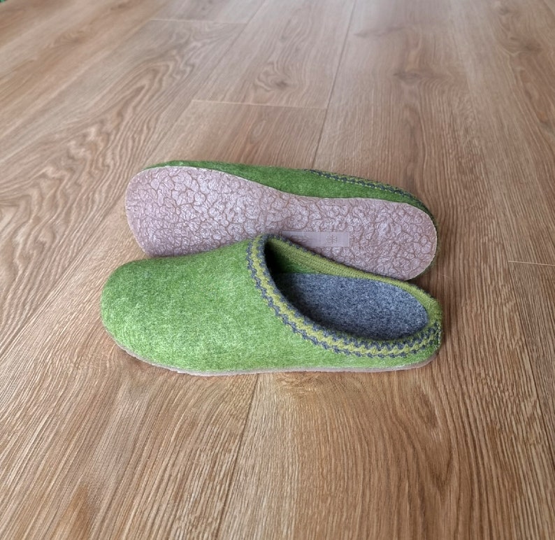 Puede incluir: Un par de zapatillas de fieltro verde con interiores y suelas grises. Las zapatillas tienen un ribete cosido en un color contrastante. Una zapatilla est&aacute; en posici&oacute;n vertical y la otra al rev&eacute;s, mostrando la suela.
