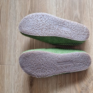Puede incluir: Dos zapatillas de fieltro verde con suelas de goma marr&oacute;n se muestran sobre una superficie de madera. Las suelas tienen un patr&oacute;n texturizado y agrietado. Las zapatillas est&aacute;n dise&ntilde;adas para uso en interiores y brindan comodidad y calidez.