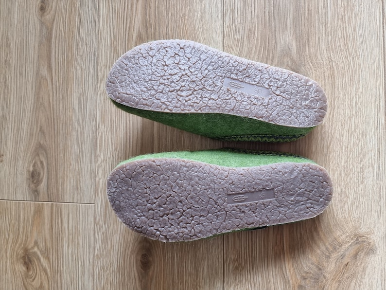 Puede incluir: Dos zapatillas de fieltro verde con suelas de goma marr&oacute;n se muestran sobre una superficie de madera. Las suelas tienen un patr&oacute;n texturizado y agrietado. Las zapatillas est&aacute;n dise&ntilde;adas para uso en interiores y brindan comodidad y calidez.