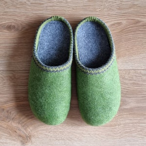 Puede incluir: Un par de zapatillas de fieltro verde con interiores grises y ribetes decorativos. Las zapatillas est&aacute;n hechas de un material suave y texturizado y est&aacute;n dise&ntilde;adas para uso en interiores. Se muestran sobre una superficie de madera.
