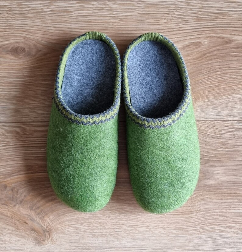 Puede incluir: Un par de zapatillas de fieltro verde con interiores grises y ribetes decorativos. Las zapatillas est&aacute;n hechas de un material suave y texturizado y est&aacute;n dise&ntilde;adas para uso en interiores. Se muestran sobre una superficie de madera.