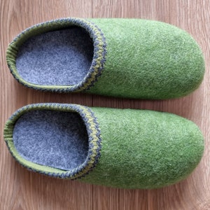 Puede incluir: Un par de zapatillas de fieltro verde. Las zapatillas tienen un interior gris y un ribete verde y gris alrededor de la abertura. Las zapatillas est&aacute;n hechas de un material suave y texturizado y est&aacute;n dise&ntilde;adas para uso en interiores.