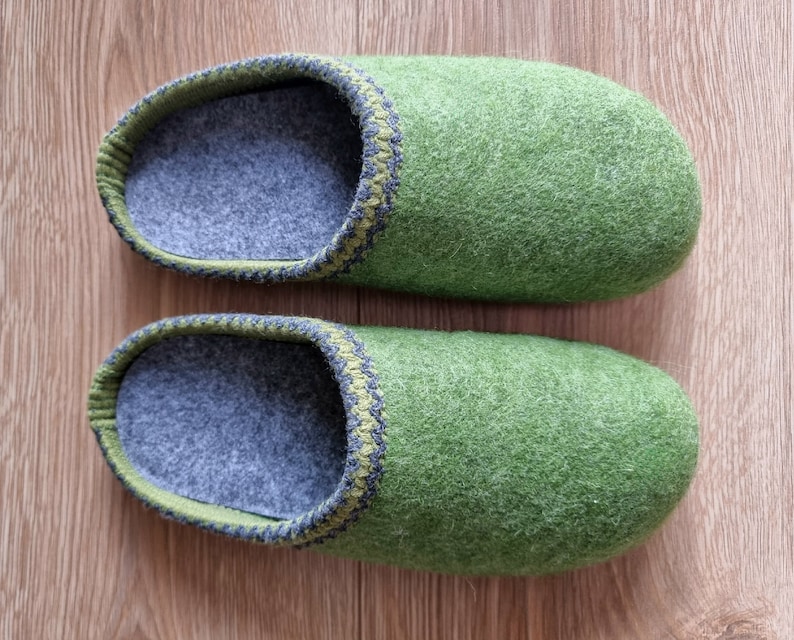 Puede incluir: Un par de zapatillas de fieltro verde. Las zapatillas tienen un interior gris y un ribete verde y gris alrededor de la abertura. Las zapatillas est&aacute;n hechas de un material suave y texturizado y est&aacute;n dise&ntilde;adas para uso en interiores.