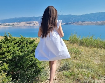 Puede incluir: Una persona joven con un vestido blanco camina por un sendero con vistas a una masa de agua. La persona tiene el pelo largo y oscuro y lleva un vestido blanco con un escote de volantes. El sendero está bordeado de arbustos verdes y el agua es de un azul brillante.