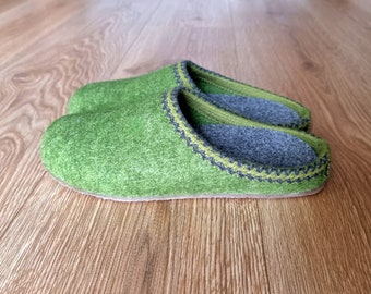 May include: A pair of green felt slippers with a gray interior and a decorative trim. The slippers are made of a soft, textured material and are designed for indoor use. The slippers are shown on a wooden surface.
