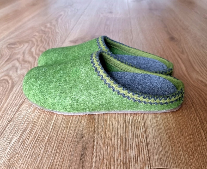 Puede incluir: Un par de zapatillas de fieltro verde con interior gris y ribete decorativo. Las zapatillas est&aacute;n hechas de un material suave y texturizado y est&aacute;n dise&ntilde;adas para uso en interiores. Se muestran sobre una superficie de madera.