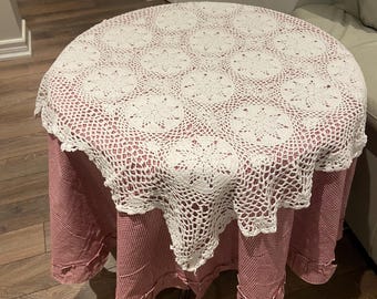 May include: A white crocheted tablecloth with a scalloped edge, placed over a red and white gingham tablecloth on a round table.