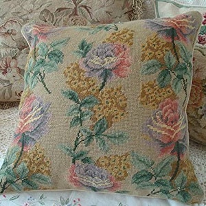 May include: A beige throw pillow with a floral pattern featuring pink, purple, and green roses. The pillow is made of a textured fabric and has a white border.