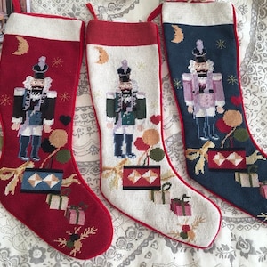 Pode incluir: Três meias de Natal com fundo vermelho, branco e azul. Cada meia apresenta um design de quebra-nozes com um esquema de cores diferente. As meias são decoradas com laços, fitas e outros detalhes festivos.