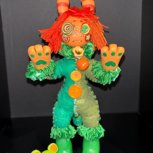 May include: A handmade doll with a whimsical design. The doll features a green and orange color scheme, with orange hair, ears, and facial details. It has green hands with paw prints, a green outfit, and green boots. A small, green bear-shaped accessory is also present.