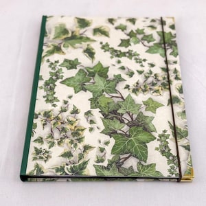 May include: A rectangular notebook featuring a cream-colored cover with a repeating pattern of green and variegated ivy leaves. The spine is solid dark green, and a thin, dark brown elastic band wraps vertically around the cover to keep it closed.