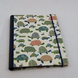 May include: A rectangular notebook with a cream-colored cover featuring a repeating pattern of small turtles in various colors and geometric designs. The notebook has a dark blue spine and a thin, vertical dark blue elastic band closure.