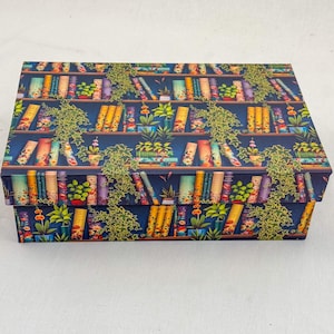 May include: Rectangular storage box with a colorful bookshelf design. The box features a dark blue background with illustrations of books, plants, and shelves. Green vines are draped across the design, adding a natural element to the decorative box.