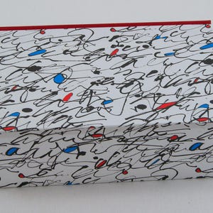 Puede incluir: Caja rectangular blanca con borde rojo y diseño garabateado en negro, rojo y azul. La caja, con tapa, es probablemente para almacenamiento o regalo.