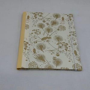 Könnte beinhalten: Eine rechteckige Mappe mit cremefarbenem Rücken und weißem Einband, der mit einem sich wiederholenden goldenen botanischen Muster aus Löwenzahn-Samenständen und Stängeln bedruckt ist. Ein dünnes weißes Gummiband umschließt den Einband vertikal.