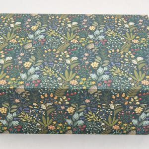 Puede incluir: Una caja rectangular con un fondo verde oscuro y un estampado floral. El patrón incluye varias flores y hojas en tonos de amarillo, rojo y verde. La caja está cerrada y se encuentra sobre una superficie blanca.