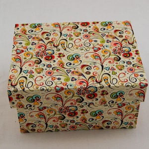 Può includere: Una scatola regalo rettangolare con sfondo color crema e un motivo floreale colorato. Il design include vortici, fiori e foglie in rosso, giallo, blu e verde. La scatola è chiusa e poggia su una superficie bianca.