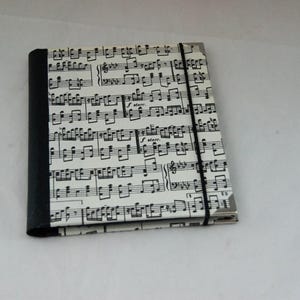 May include: A rectangular notebook featuring a cover with a black and white musical score print. The spine is solid black, and a thin black elastic band wraps vertically around the cover to keep it closed. Metal corner protectors are visible.
