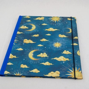 May include: A rectangular folder with a dark blue cover featuring a repeating pattern of golden suns, crescent moons, and clouds. The folder has a solid blue spine and a thin black elastic band stretched vertically to keep the cover closed.