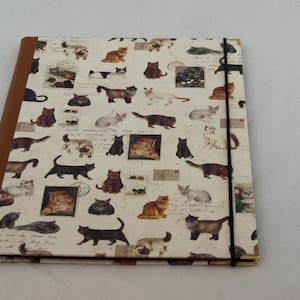 May include: A brown and cream binder with a black elastic closure. The binder is covered in a pattern of cats in various poses and colours.