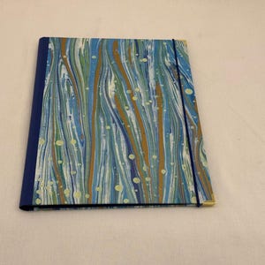 Può includere: Un taccuino chiuso con un design marmorizzato nei toni del blu, verde e oro. Il taccuino ha un dorso blu scuro e una chiusura con elastico abbinato. La copertina presenta un motivo astratto e fluido.