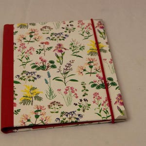 Puede incluir: Una carpeta blanca con lomo rojo y un patrón floral. La carpeta tiene un cierre de banda elástica roja.
