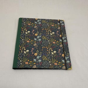 Könnte beinhalten: Ein quadratisches Notizbuch mit dunklem floralem Muster auf dem Einband mit einer Vielzahl von Blumen und Blättern in Gelb-, Blau- und Orangetönen. Der Buchrücken ist grün, und ein schwarzes Gummiband sichert das Notizbuch. Die Ecken sind goldfarben.
