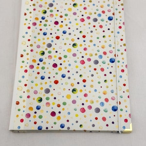 May include: A rectangular notebook with a white cover featuring a pattern of multicolored watercolor-style dots in shades of blue, yellow, pink, green, and purple. The notebook includes a white elastic closure band and gold-toned metal corner protectors.