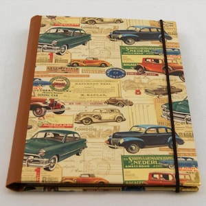 May include: A vintage-style notebook with a brown leather cover and a black elastic band closure. The cover features a collage of vintage cars, travel posters, and luggage labels. The text on the cover includes "Morecambe", "London", "Baggage Room", "Station Hotel", "Southern Railway", "Dining Car", "Railroad Pass", "M. Kaplan", "New York", "California", "Amsterdam", and "Nederland".
