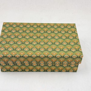 Puede incluir: Caja rectangular cerrada con un patrón repetitivo en forma de abanico en verde, naranja y blanco. El patrón cubre la parte superior y los lados de la caja, que descansa sobre una superficie blanca.