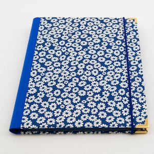 May include: A square notebook with a blue cover and a white daisy pattern. The spine is solid blue, and a blue elastic band secures the notebook. Gold-colored corner protectors add a touch of elegance.