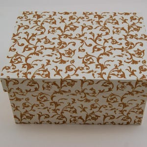 Puede incluir: Una caja rectangular con tapa, con un fondo blanco y un patrón repetido de diseños florales y de desplazamiento de color dorado. La caja probablemente está hecha de cartón o papel.