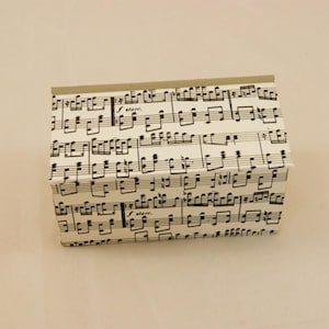 Puede incluir: Una pequeña caja rectangular con fondo blanco y notas musicales negras impresas en la superficie. La caja es de cartón y tiene una tapa con bisagras.