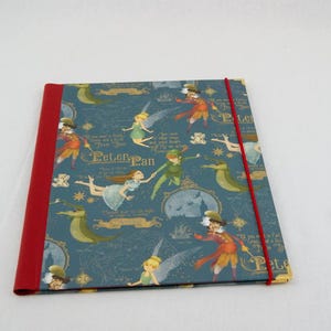 May include: A rectangular notebook with a dark blue cover featuring a repeating Peter Pan pattern, including illustrations of Peter Pan, Wendy, Tinker Bell, and Captain Hook. The notebook has a solid red spine and a matching red elastic closure band.
