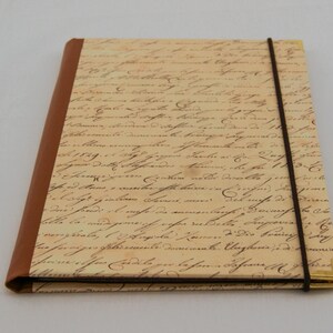 May include: A rectangular notebook featuring a cream-colored cover with a vintage-style cursive script print. The spine is bound in brown material, and a thin dark brown elastic band wraps around the cover. Gold-toned metal corners protect the edges.