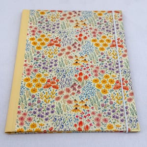 May include: A rectangular notebook featuring a cream-colored cover with a dense, colorful watercolor-style floral pattern of red poppies, yellow daisies, purple lavender, and blue wildflowers, finished with a yellow spine and a white elastic band.