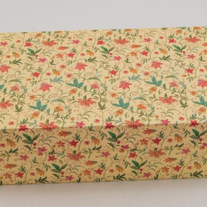 Puede incluir: Una caja rectangular con un estampado floral. La caja está cubierta con un diseño repetido de flores rojas, naranjas y rosas con hojas verdes sobre un fondo amarillo claro. La caja está cerrada.