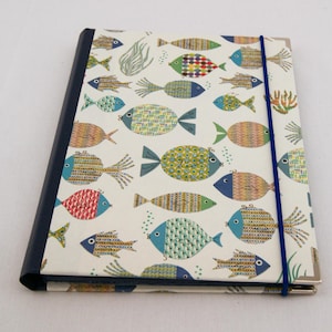 May include: A rectangular notebook with a white cover featuring a repeating pattern of multi-colored fish in shades of blue, green, yellow, and red. The notebook has a dark blue spine and a thin blue elastic band closure with silver metal corners.