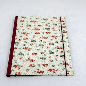 May include: A rectangular portfolio folder featuring a cream-colored cover with a repeating pattern of small red and green floral clusters. The folder has a solid dark red spine and a matching dark red elastic band closure with gold corner accents.