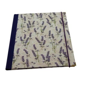 May include: A rectangular notebook featuring a cream-colored cover with a repeating pattern of purple lavender sprigs and faint cursive script text. The spine is bound in solid dark purple fabric, and a thin purple elastic band secures the cover.