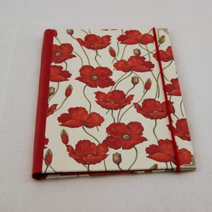 May include: A rectangular notebook featuring a cream-colored cover with a repeating pattern of red poppy flowers, buds, and thin green stems. The notebook has a solid red spine and a vertical red elastic band closure attached to the right side.