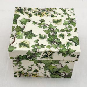 Pode incluir: Uma caixa quadrada com tampa apresentando um padrão repetitivo de folhas e vinhas de hera verde sobre um fundo creme. O design cobre o topo e as laterais da caixa, exibindo ilustrações detalhadas de várias formas de folhas de hera.
