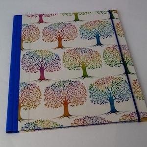 May include: A square notebook with a white cover featuring a repeating pattern of colorful trees. The trees have rainbow-colored leaves and brown trunks. The spine is blue, and a blue elastic band secures the notebook.