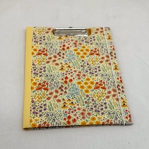May include: A clipboard with a floral pattern and a metal clip. The design features a cream-coloured background with a colourful floral pattern in shades of red, yellow, blue, and purple. It has a yellow spine and a white elastic band.